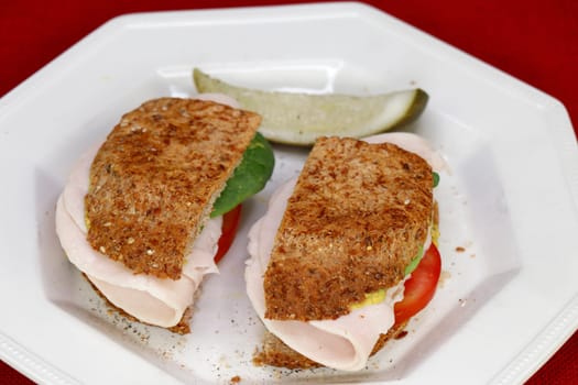 Close-up of two halves of a turkey sandwich with tomato, spinach, mustard and ground black pepper with a dill pickle spear on a plate. Homemade turkey sandwich with a dill pickle spear on a plate