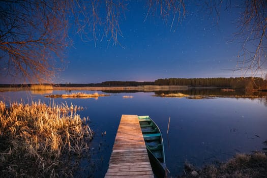 Belarus, Eastern Europe. Real Night Sky Stars Above Old Pier With Moored Wooden Fishing Boat. Natural Starry Sky And Countryside Landscape With Lake River In Early Spring Night. Russian Nature.