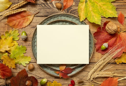 Autumn rustic table setting with blank card between colorful leaves and berries, top view. Horizontal card mockup on wooden table, copy space. Thanksgiving, halloween or harvest concept