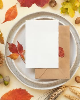 Autumn table place with colorful leaves and blank card with envelope top view. Mockup of fall vertical card on light yellowtable, flat lay, copy space. Harvest, Halloween and Thanksgiving season