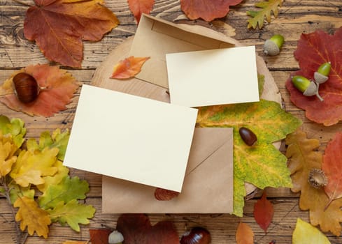 Blank cards between colorful autumn leaves and berries on wooden table top view, rustic mockup. Horizontal cards over envelopes, flat lay, copy space. Thanksgiving, halloween or harvest concept