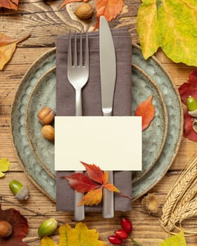 Autumn rustic table place with blank card between colorful leaves and berries, top view, mockup. Flat lay with horizontal card on wooden table, copy space. Thanksgiving or harvest concept