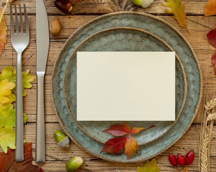 Autumn rustic table setting with blank card between colorful leaves and berries, top view. Horizontal card mockup on wooden table, copy space. Thanksgiving, halloween or harvest concept