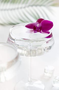 Transparent cocktail with ice in a champagne coupe glass decorated with purple orchid flowers close up. Tropical drink at the bar. Summer vacation mood