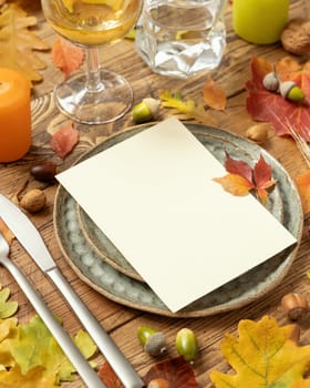 Autumn rustic table setting with blank card between colorful leaves and berries, close up Vertical card mockup on wooden table, copy space. Thanksgiving, halloween or harvest concept