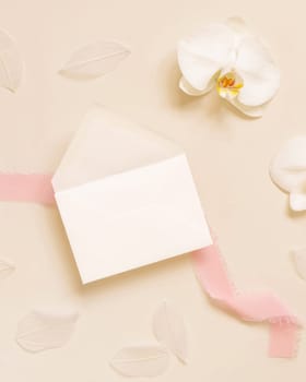 Wedding envelope near white orchid flower and silk ribbons on light yellow top view, mockup. Tropical scene with horizontal blank card flat lay
