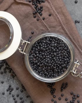 Glass jar of dry black lentils beans on grey table top view. Healthy eating, protein source for vegan and vegetarian diet. Traditional Asian and North African cuisines ingredient
