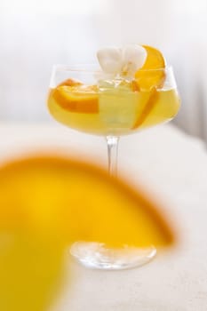 Transparent cocktail with ice cubes in a champagne coupe glass decorated with slices of orange and white orchid flower close up. Tropical drink at the bar. Summer vacation mood
