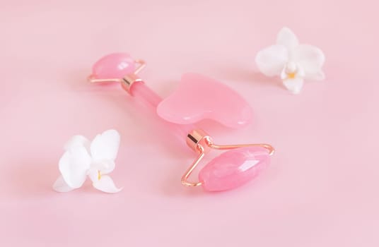 Rose quartz facial roller and gua sha massager near white orchid flowers, close up on light pink. Anti-age massage and natural lifting therapy at home. Facial skin care at home. Beauty concept