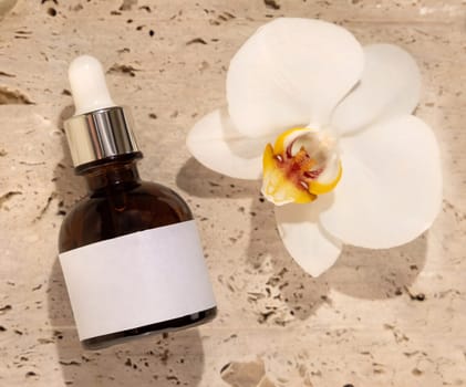 Brown Glass Dropper bottle on stone near white orchid flower top view, label mockup. Skincare handmade beauty product, serum or lotion. Exotic natural cosmetics..