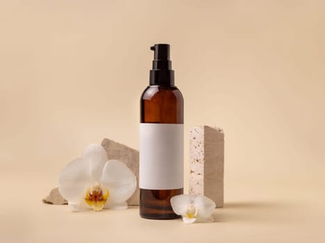 Dark glass cosmetic bottle with blank label near white orchid flowers and stones on light yellow, close up, mockup. Skincare handmade beauty product, tonic or lotion. Exotic natural cosmetics..
