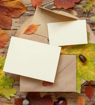 Blank cards between colorful autumn leaves and berries on wooden table top view, rustic mockup. Horizontal cards over envelopes, flat lay, copy space. Thanksgiving, halloween or harvest concept