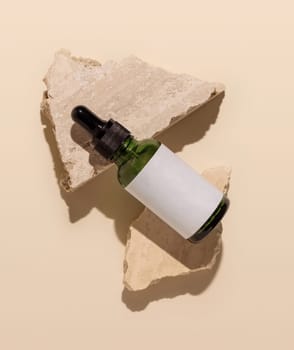 Green glass dropper bottle lies on beige travertine stones on light yellow top view, mockup. Skincare beauty product package. Natural cosmetics, minimal composition, hard shadows ..