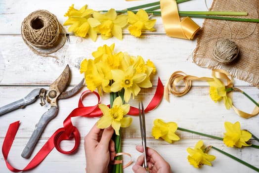 Top view of florist hands making flower bouquet with narcissus and decorative ribbon.