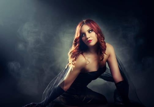 Beautiful red hair woman with evening makeup, glamour hairstyle and elegant shiny dress in front of smoke and studio lights.