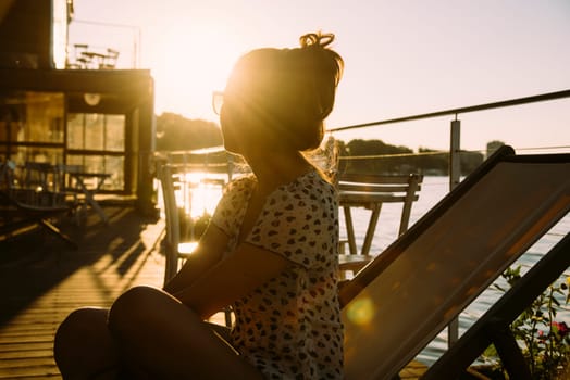 Young hipster woman in summer clothing sitting in cafe, enjoys the sun and watching the river at summer sunset. Retro style image.