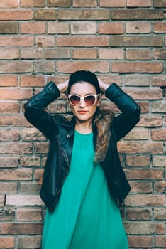 Fashionable young woman in leather jacket and sunglasses in front of brick wall background