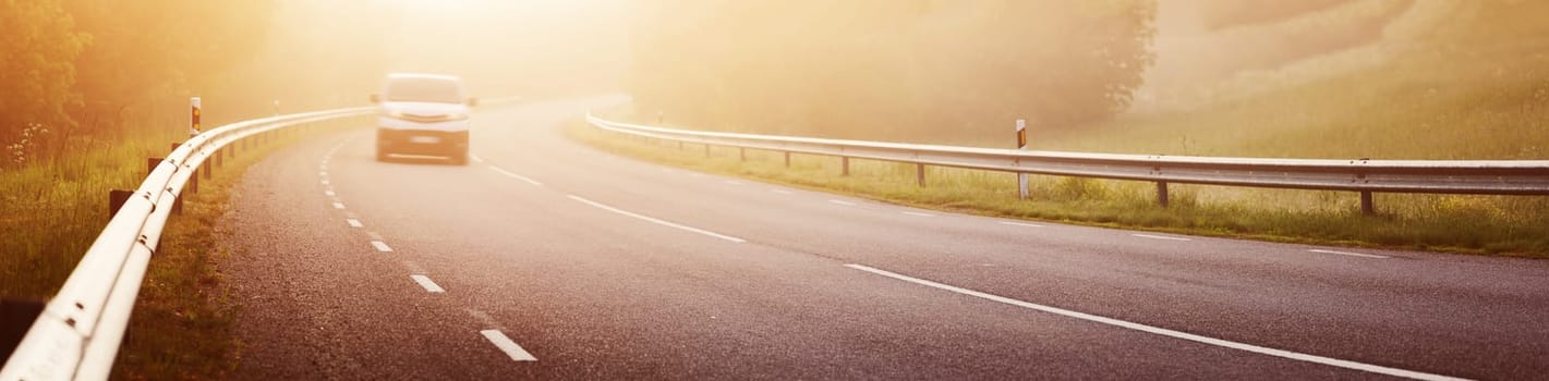 Car on the asphalt road with dividing line and safety fence in the morning mist. Highway in sunrise in natural park.