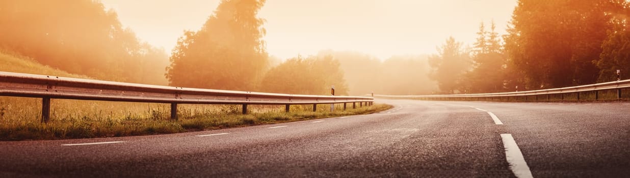 Black asphalt road with dividing line and safety fence in the morning mist. Highway in sunrise in natural park.