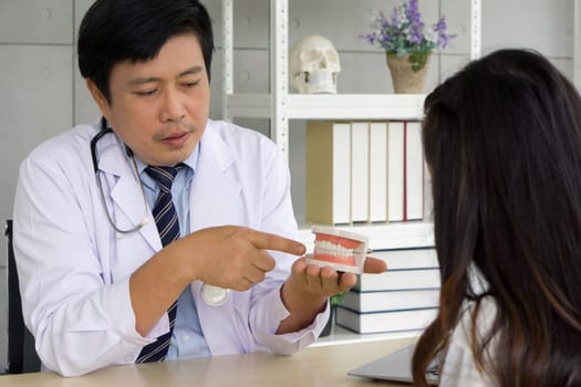 Asian dentist in white gown and stethoscope pointing finger at tooth model, explain to young patient about orthodontics and braces. Help reduce gum pain. Healthcare and medicine concept.