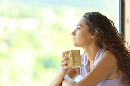 Side view portrait of a woman holding coffe cup relaxing looking through a window