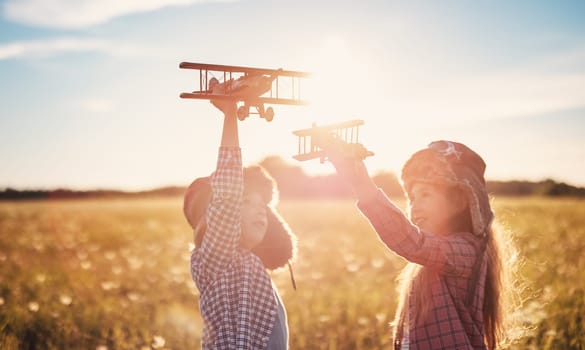 Boy and girl plaing with wooden planes on the field in sunset. Concept of the family vacation, dreaming and friendship.