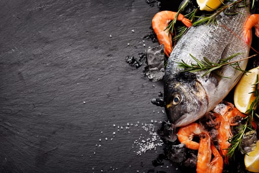 Top view of fresh sea food on grunge textured black background. Shrimps, bream, herbs, lemon and ice on slate with copyspace.