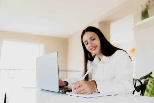 Caucasian business woman sitting at desk, reading from computer, taking notes at notebook. Female student using pc, writing down important information with pencil in note pad. Happy smiling lady