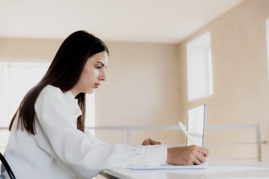 Focused Caucasian young business woman sitting at desk, reading from computer, taking notes at her white notebook. Female student using pc, writing down important information with pencil in note pad