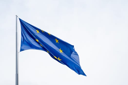 Flag of the European Union waving from a flagpole. Against the blue background, the golden stars form a circle as a symbol of unity and solidarity among the peoples of Europe. The number of stars on the flag is invariable and does not depend on the number of member countries. Horizontal composition and copy space for text.