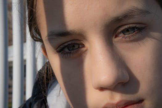Human face cropped in an extreme close-up of a teenage girl staring tenderly at the camera. Sunlight illuminates her face, her eyes are green and one of them is partially covered by a strip of shadow.