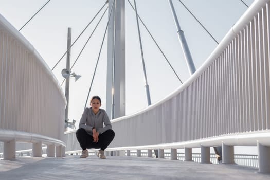 Attractive teenage girl in sportswear sitting on her haunches along the pedestrian walkway of a cable-stayed bridge, looking at camera with serious face expression. Horizontal and full length composition.