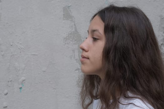 Close up profile of a teenage girl with long brown hair looks forward with a thoughtful expression. Background of gray wall, copy free space for text.