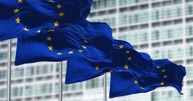 group of flags of the European Union waving in the wind in front of the European Parliament building.