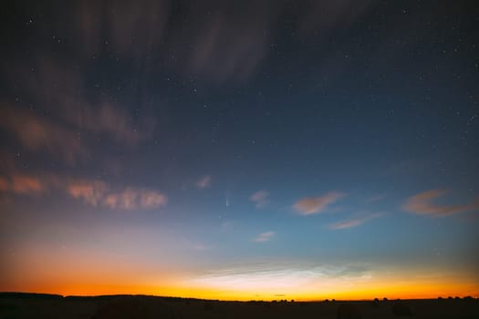 Night Starry Sky With Glowing Stars Above Countryside Landscape. Light Cloudiness Overcast Above Rural Field Meadow In Early Spring. Illumination Lights Of Town On Horizon.