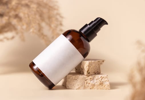 Brown glass cosmetic bottle with blank label on stones near pampas grass on light yellow, close up, mockup. Skincare beauty product. Natural eco friendly cosmetics
