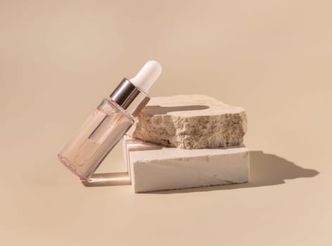 Glass cosmetic dropper bottle on beige travertine stones on light yellow close up, mockup. Skincare natural beauty product. Minimal eco friendly composition.