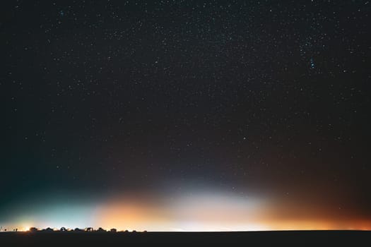Night Starry Sky With Glowing Stars Above Countryside Town Landscape. Rural Field Meadow In Early Spring. Bright Glow Of Stars. Illumination Lights Of Small Town Or Village.