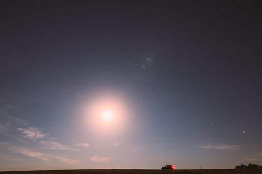 Full Moon Rising Above Field In Countryside. Night View Of Natural Glowing Stars. Car Parked On Field Under Blue Night Starry Sky With Moon.