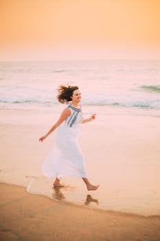 Goa, India. Young Caucasian Woman With Fluttering Hair In Wind In White Dress Walking Along Seashore, Enjoying Life And Smiling In Summer Sunlight.