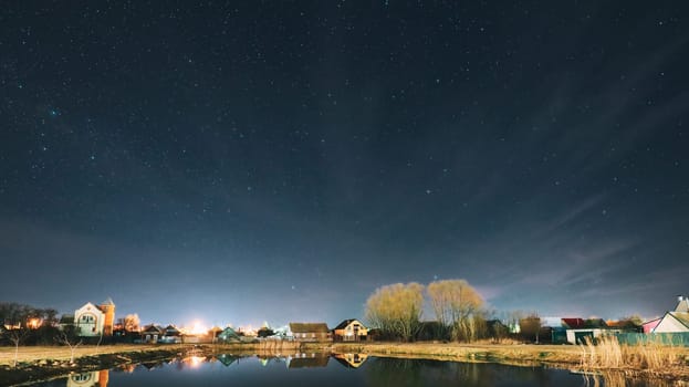 Belarus, Eastern Europe. Night Sky Stars Above Countryside Landscape With Lake Coast And Small Town Or Village. Natural Starry Sky Above Pond And Houses In Early Spring Night. Panorama Panoramic View