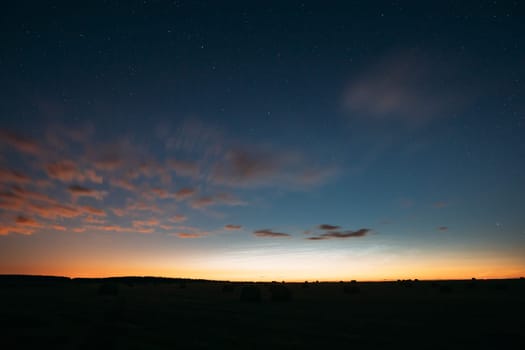 Night Starry Sky With Glowing Stars Above Countryside Landscape. Light Cloudiness Overcast Above Rural Field Meadow In Early Spring. Illumination Lights Of Town On Horizon.