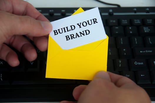 Hand holding with notepad in yellow envelope with text - Build your brand. With keyboard monitor background. Branding concept.