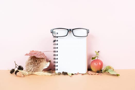 Empty notebook with eyeglasses, an apple and a hedgehog, autumn template with colorful leaves, office desk, pastel color