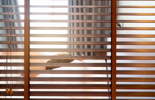Wooden Venetian blinds. Blackout curtain between bathroom and bedroom in hotel with back view of woman sleep under white blanket on bed in the morning. Jalousie or window blinds. Horizontal blinds.