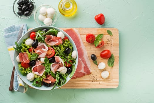 Healthy green salad with prosciutto, mozzarella, tomato and olives on green background