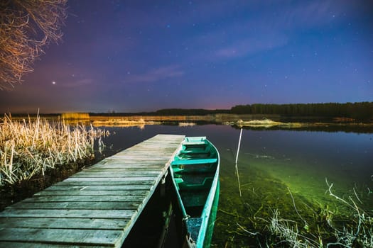 Real Night Sky Stars Above Old Pier With Moored Wooden Fishing Boat. Natural Starry Sky And Countryside Landscape With Lake River In Early Spring Night. Russian Nature.