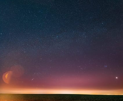 Night Starry Sky With Glowing Stars Above Countryside Landscape. Milky Way Galaxy And Rural Field Meadow In Early Spring. Bright Glow Of Planet Venus Among The Stars. Warm Lights Of Evening Sunset.