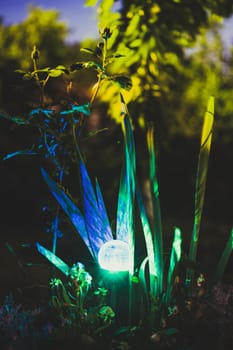 Night View Of Flowerbed Illuminated By Energy-Saving Solar Powered Colorful Multi-colored Lantern On Yard. Beautiful Small Garden With Blue And Green Light, Lamp In Flower Bed. Garden Design.