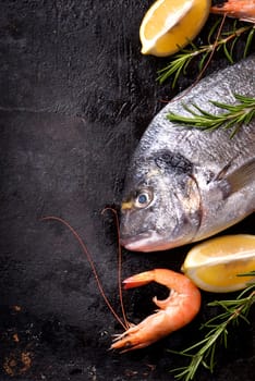 Top view of fresh sea food on grunge textured black background. Shrimps, bream, herbs,and lemon on slate with copyspace.